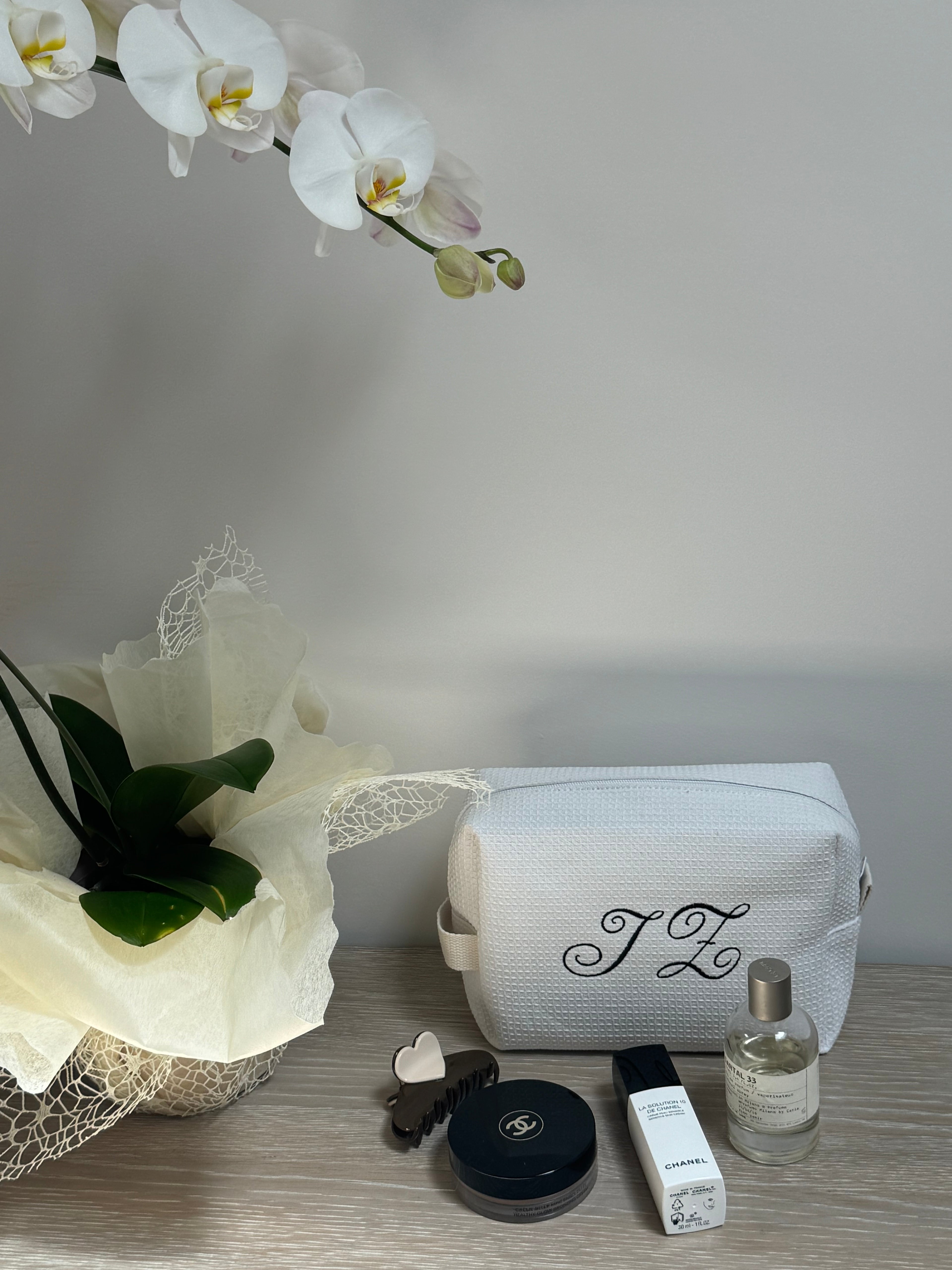 White textured toiletry bag with custom initials, displayed with luxury beauty products and a potted orchid.