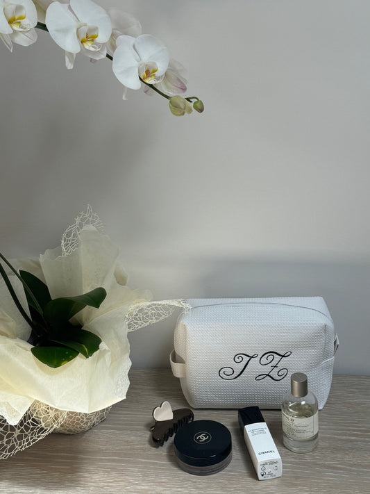 White textured toiletry bag with custom initials, displayed with luxury beauty products and a potted orchid.